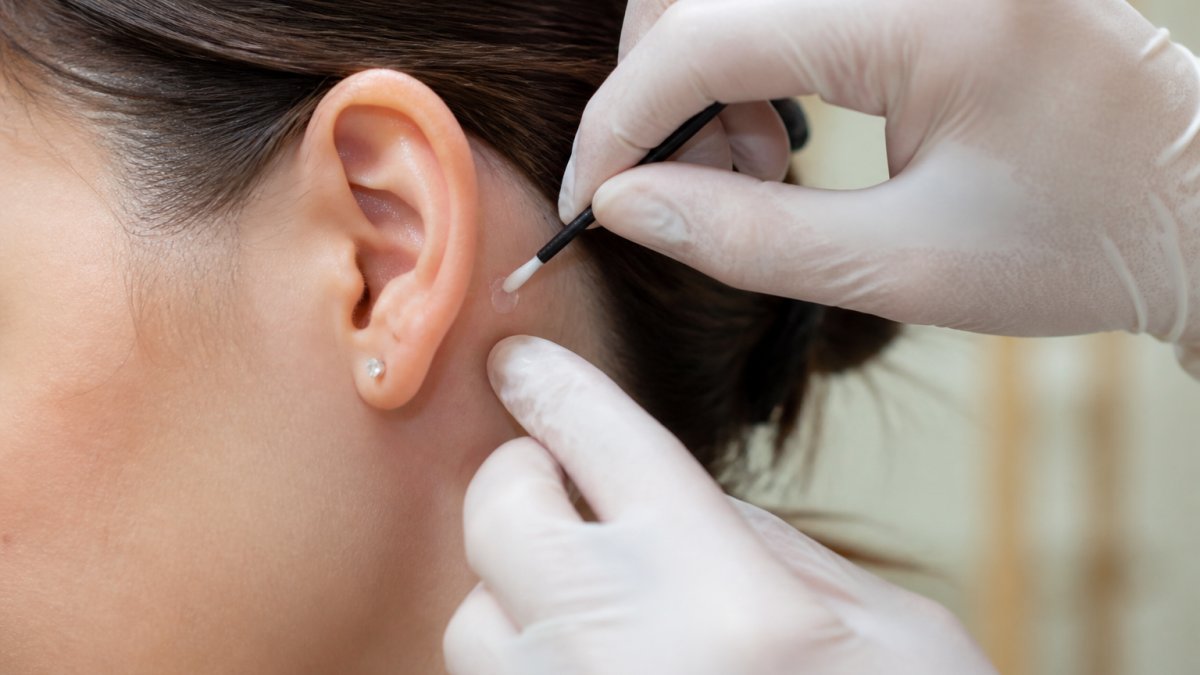 Close-up of a lash artist's gloved hands applying a small drop of clear adhesive to a client's clean skin behind the ear with a microbrush, bright professional salon lighting, soft neutral background